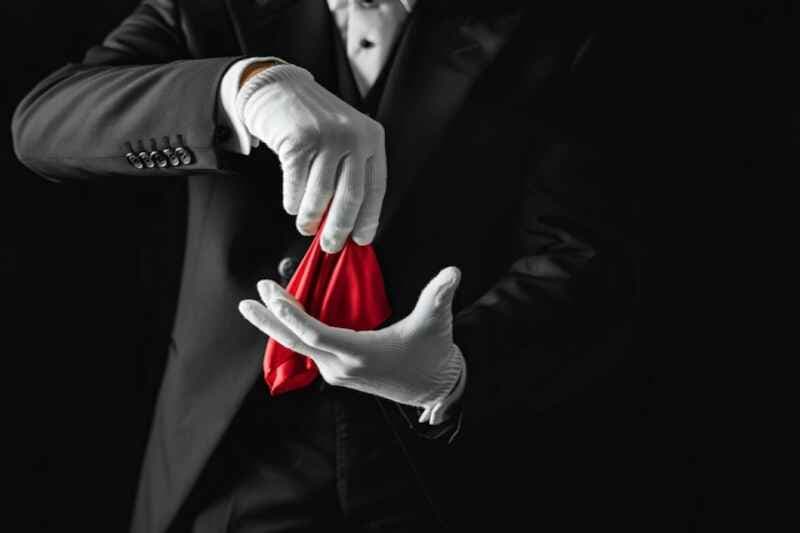 A magician wearing a tuxedo and white gloves holds a red object, performing a mysterious illusion against a dark background.