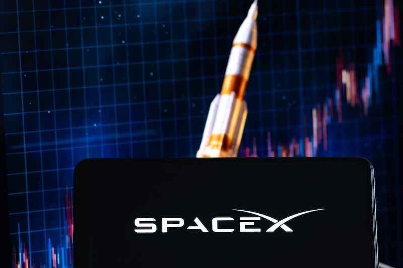 SpaceX logo displayed on a smartphone screen against a background of a rising stock chart and a rocket shown on a computer monitor, illustrating the concept of investment and the debut of Elon Musk’s company SpaceX
