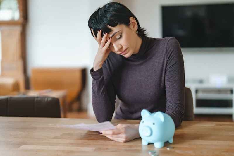 Woman looks worried while reading a bill and sitting at a table with a piggy bank at home